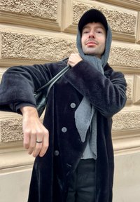 Manteau en peluche noir avec de gros boutons, écharpe grise enroulée autour du cou, et un sac à main noir. En arrière-plan, un mur beige texturé.