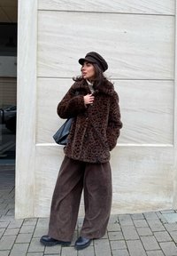 Manteau en fausse fourrure imprimé léopard marron avec un large col, fermeture à boutons ; assorti à un pantalon large marron foncé et des bottines noires. Sac cabas noir.