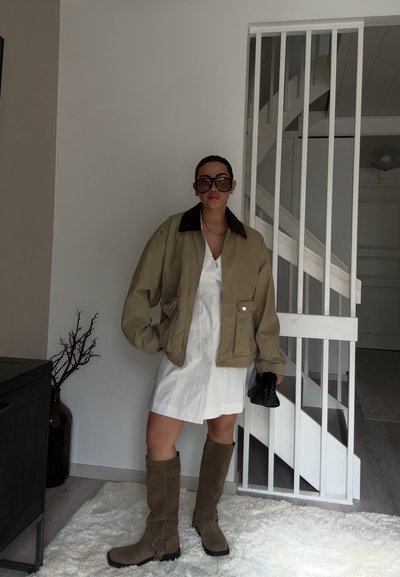 Chaqueta bomber de algodón color caqui con un cuello marrón, combinada con un vestido blanco y botas marrones hasta la rodilla. Los accesorios incluyen un bolso negro.