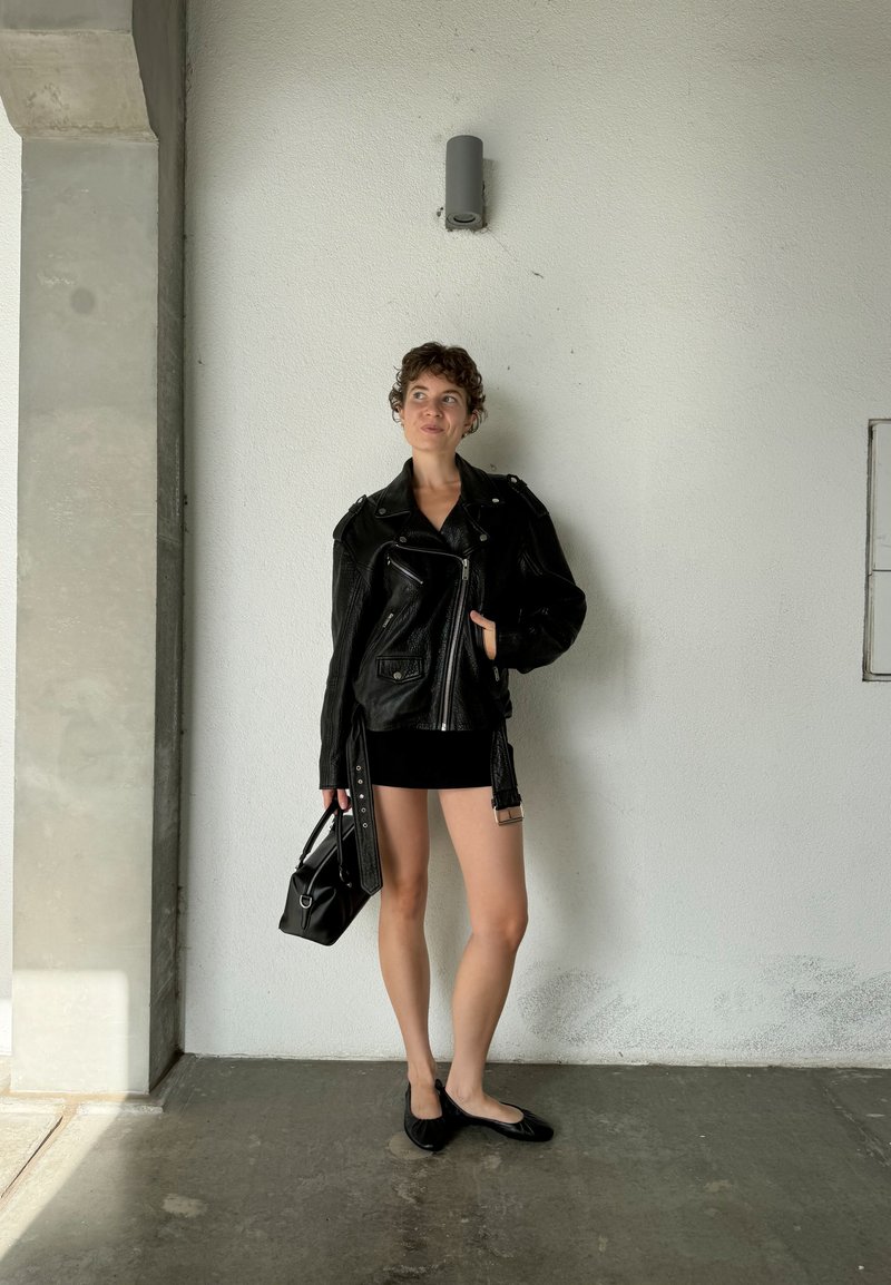 Casaco de motoqueiro de couro preto com acabamento texturizado, sobre um vestido mini preto ajustado. Uma bolsa preta e sapatos rasos completam o visual.
