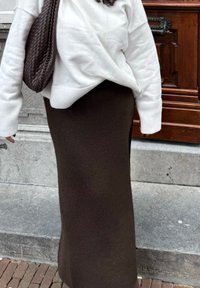 Adīta brūna maksi svārki, kas apvienoti ar baltu svīteri, ar ribotām piedurknēm un teksturētu brūnu austu rokassomu.