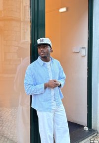 Chemise à rayures bleues et blanches à boutons, sous-vêtement blanc, pantalon blanc à jambes larges, casquette grise avec logo, debout près d'une porte en verre.