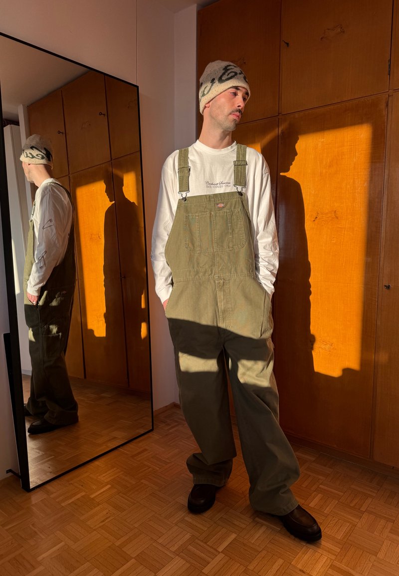 Hombre con gorro de punto beige, camisa blanca de manga larga y overoles verde oliva junto a un espejo de cuerpo entero en una habitación iluminada por el sol.