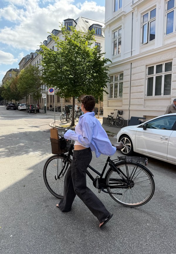 Persoon die met een zwarte fiets wandelt op een straat in een zonnig stedelijk gebied met geparkeerde auto's en bomen langs het trottoir.