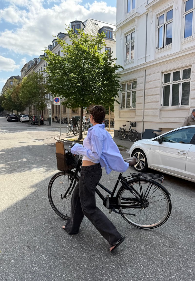 Personne marche avec un vélo noir dans une rue d'une zone urbaine ensoleillée, avec des voitures garées et des arbres le long du trottoir.