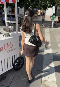 Zwarte handtas met een glanzende afwerking, witte rok met een aangesloten ontwerp en een zilveren armband. Een kopje in de hand, grijze stoeptegels en een kar in de buurt.