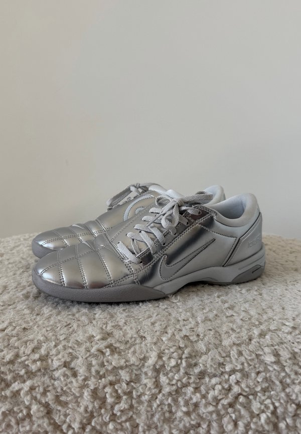 Ζευγάρι ασημένια αθλητικά παπούτσια Nike με κορδόνια, τοποθετημένα δίπλα-δίπλα σε ένα φωτεινό υφασμάτινο χαλί μπροστά σε ένα απλό φόντο τοίχου.