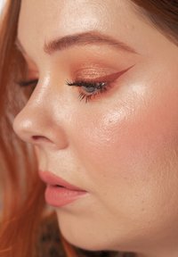 Close-up af et ansigt med varm kobberøjenskygge, markant vingeformet eyeliner og glat, strålende hudtone; med bløde læber og fremtrædende kindben.