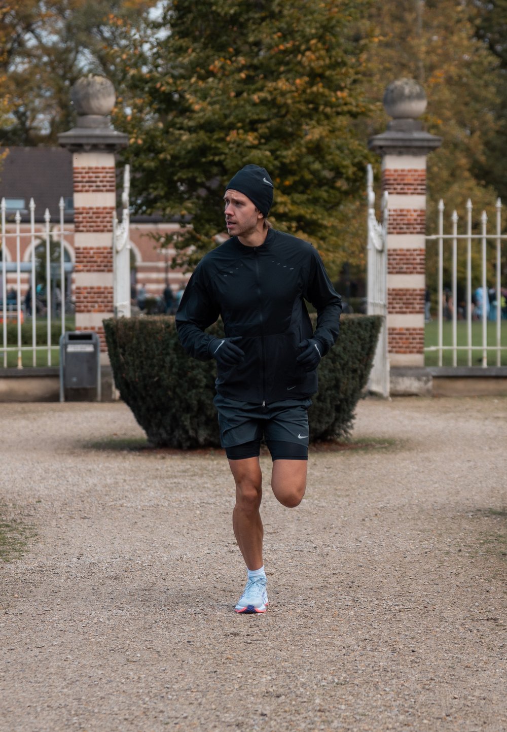 Fekete sportdzseki és rövidnadrág, kék sportcipő, fut a kavicsos úton egy parkban, háttérben zöld cserjék és fák.