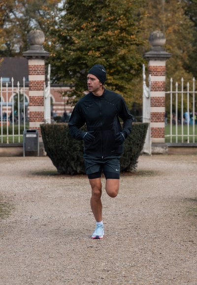 Chaqueta deportiva negra y pantalones cortos, zapatillas azules, corriendo por un camino de grava en un parque con arbustos verdes y árboles de fondo.