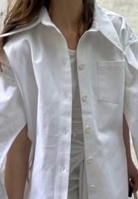 Camisa oversized branca feita de algodão, com colarinho pontudo, um bolso no peito e seis botões na parte da frente, com mangas enroladas.