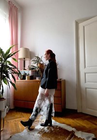 Svart blazer over en hvit blonderkjole, kombinert med blanke svarte støvler som går opp til knærne. Innendørs setting med tremøbler og planter.