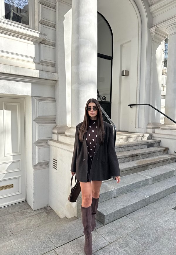 Kvinde i sort oversized blazer og polkaprik kjole går ned ad stentrapper uden for en hvid søjlebåret bygning, bærer håndtaske og solbriller.