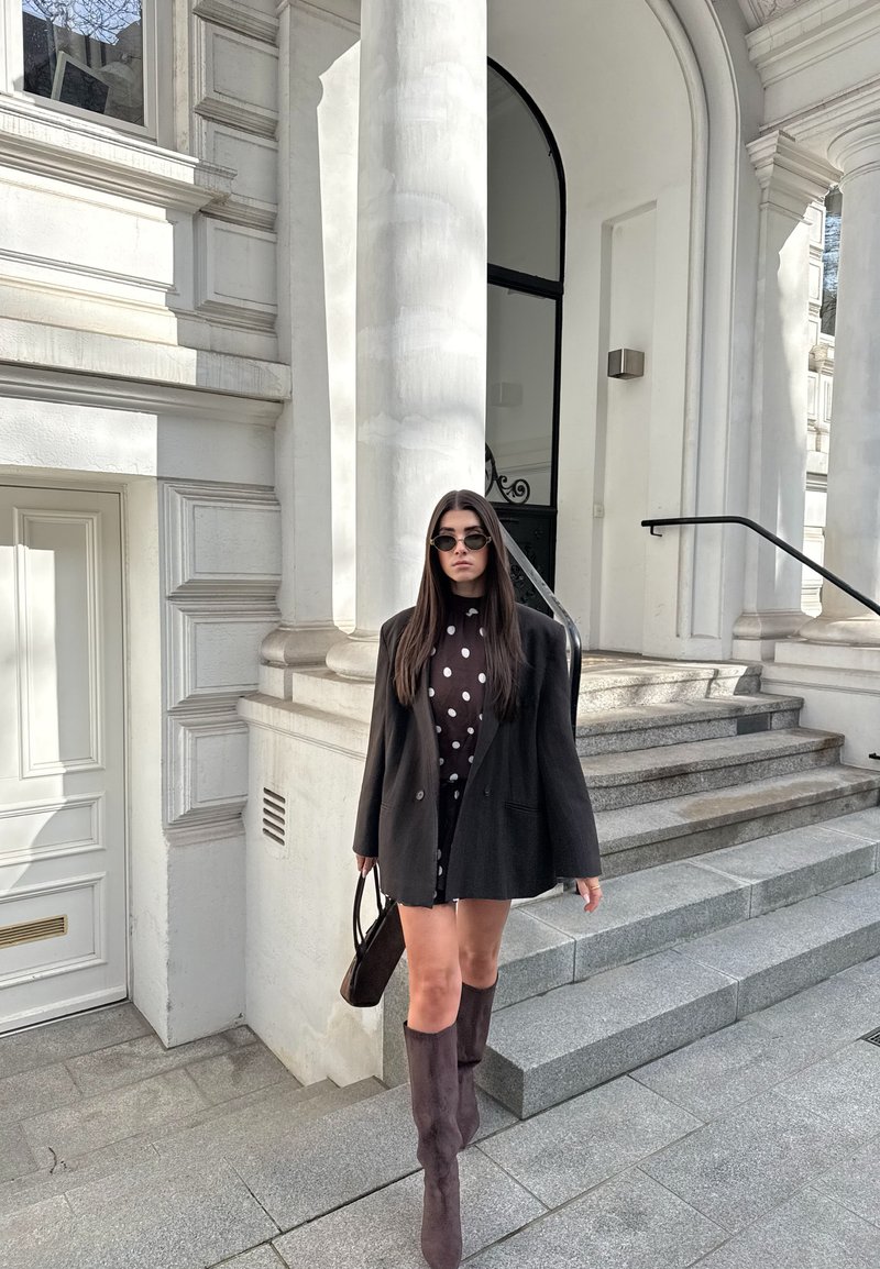 Γυναίκα με μαύρο oversized σακάκι και πουά φόρεμα κατεβαίνει πέτρινα σκαλοπάτια έξω από ένα λευκό κτίριο με κολόνες, κρατώντας τσάντα και φορά γυαλιά ηλίου.