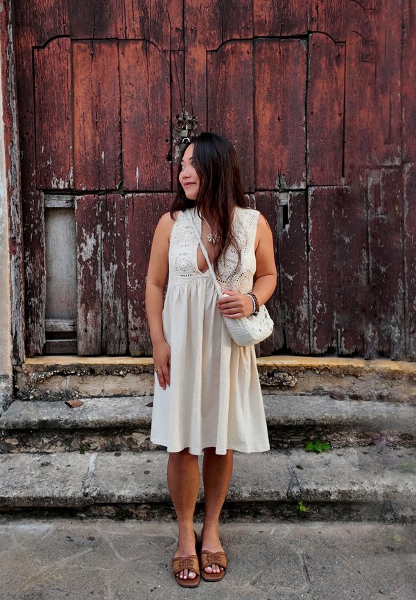 Kvinde i en cremefarvet ærmeløs kjole med en lille hvid taske, stående barfodet på stentrapper foran vejrbidte trædøre.