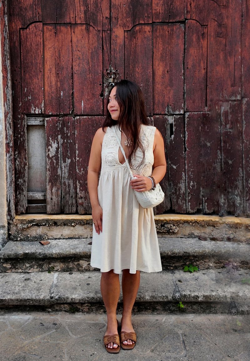 Mujer con vestido sin mangas color crema y un pequeño bolso blanco, de pie descalza sobre escalones de piedra frente a puertas de madera desgastadas.