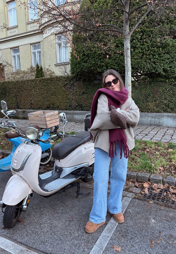 White scooter with a brown seat and chrome accents parked next to a woman wearing a grey oversized jumper, blue jeans, and a burgundy scarf.