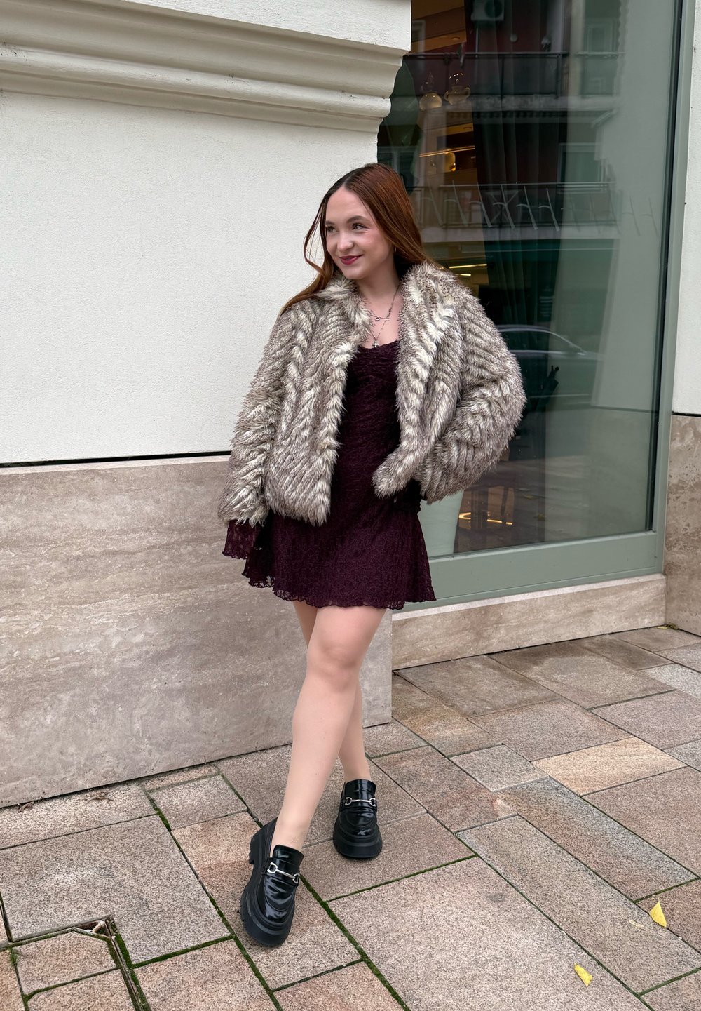 Fiatal nő hosszú hajjal, bolyhos bunda kabátban, sötét csipkés ruhában és fekete magassarkú platform cipőben áll egy városi járdán.