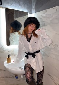 Personne portant un chapeau en fourrure noire, une chemise blanche oversize ceinturée, et des collants à motifs, appuyée sur un lavabo de salle de bain avec des murs en marbre.