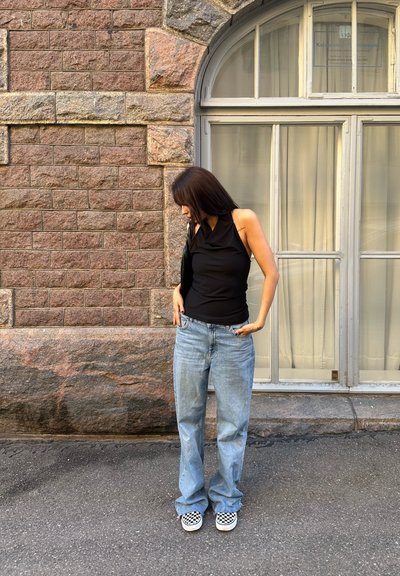 Top negro sin mangas, jeans desgastados de color azul claro y zapatos de lona de cuadros, de pie contra una pared de piedra y una gran ventana.