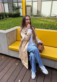 Mulher com um trench coat bege e jeans sentada num banco amarelo, a aplicar batom, segurando uma bolsa castanha, em um deck de madeira perto da relva.