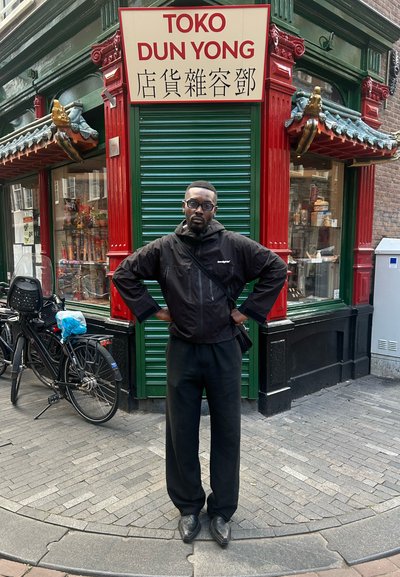 Chaqueta negra impermeable con capucha, pantalones negros, de pie frente a una fachada de tienda roja y verde con letreros en inglés y chino.