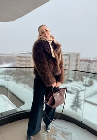 Veste en fausse fourrure marron, pull crème avec capuche, pantalon large noir et sac à main bordeaux foncé. Debout sur un balcon avec de la neige.