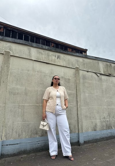 Mujer con pantalones blancos anchos, cárdigan de punto beige de manga corta, gafas de sol, sosteniendo un pequeño bolso blanco, de pie junto a una pared de hormigón desgastada.