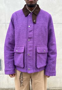 Personne portant une veste en laine violette avec un col en velours côtelé marron et des boutons-pression argentés, associée à un pantalon beige et un accessoire perlé.