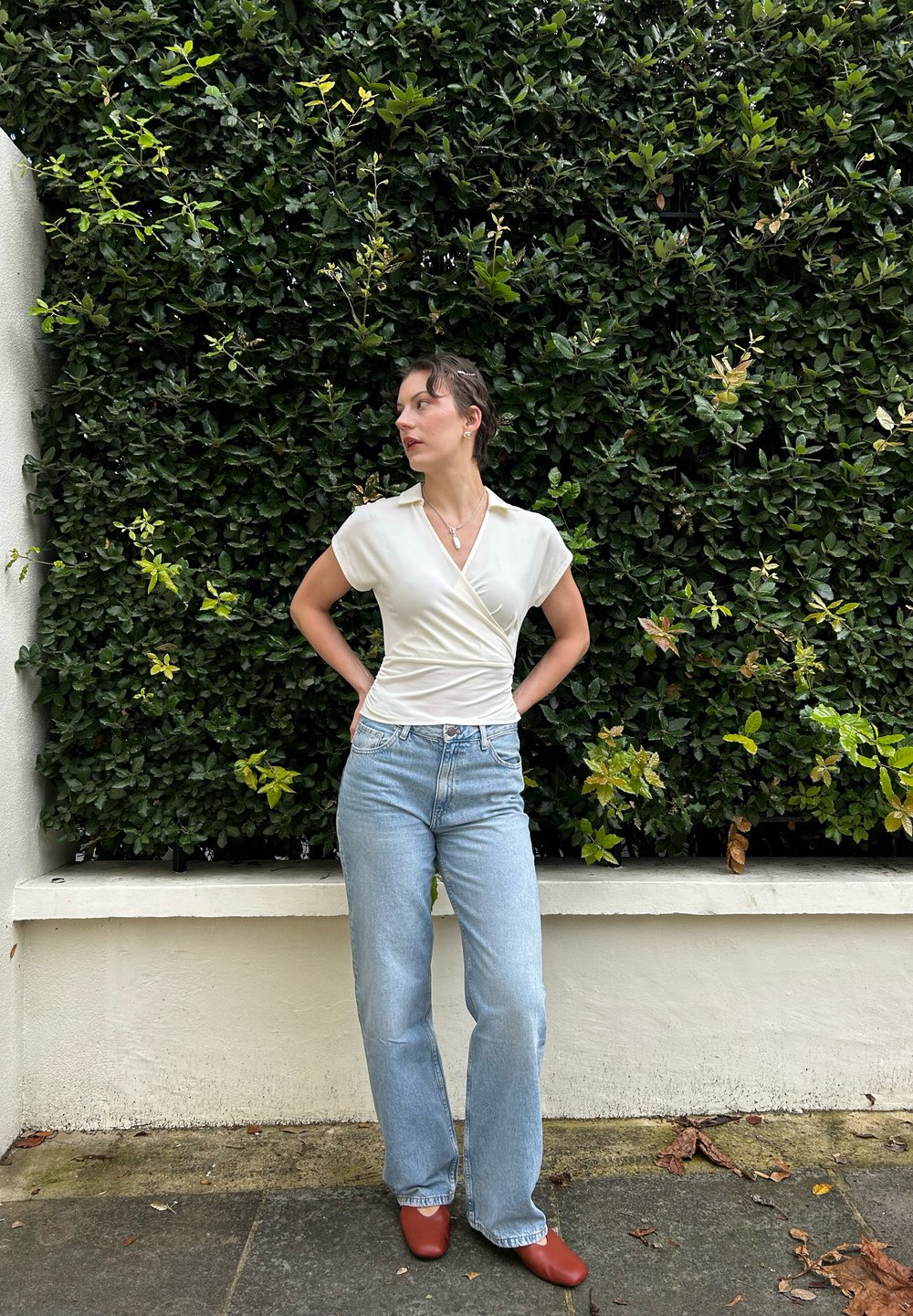 Λευκή μπλούζα τύπου wrap, ανοιχτό μπλε τζιν με ίσια γραμμή και κόκκινες παντόφλες, σε φόντο πράσινης βλάστησης. Η μπλούζα έχει γιακά; απαλό υφαντό υ�λικό.