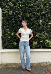 Λευκή μπλούζα τύπου wrap, ανοιχτό μπλε τζιν με ίσια γραμμή και κόκκινες παντόφλες, σε φόντο πράσινης βλάστησης. Η μπλούζα έχει γιακά; απαλό υφαντό υλικό.