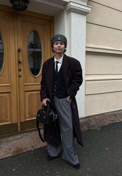 Abrigo largo oscuro, chaleco negro, camisa blanca, corbata negra, boina gris, pantalones de rayas anchas, bolso negro con acentos metálicos, de pie junto a puertas de madera.