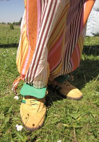 Žlté tenisky Adidas s zelenými akcentmi, spárované s multicolornými pásikavými nohavicami v oranžovej, ružovej a bielej farbe, zobrazené z nízkeho uhla na tráve.