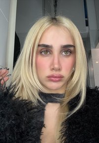 Jeune femme aux cheveux blonds raides, yeux verts, lèvres brillantes, portant une veste noire duveteuse et un haut foncé, regardant légèrement à côté de l'appareil photo.