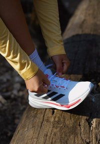 Sapatilha branca desportiva com riscas pretas, detalhes em vermelho e parte superior texturizada, sendo atada por mãos. Usada com uma t-shirt de manga longa amarelo claro.