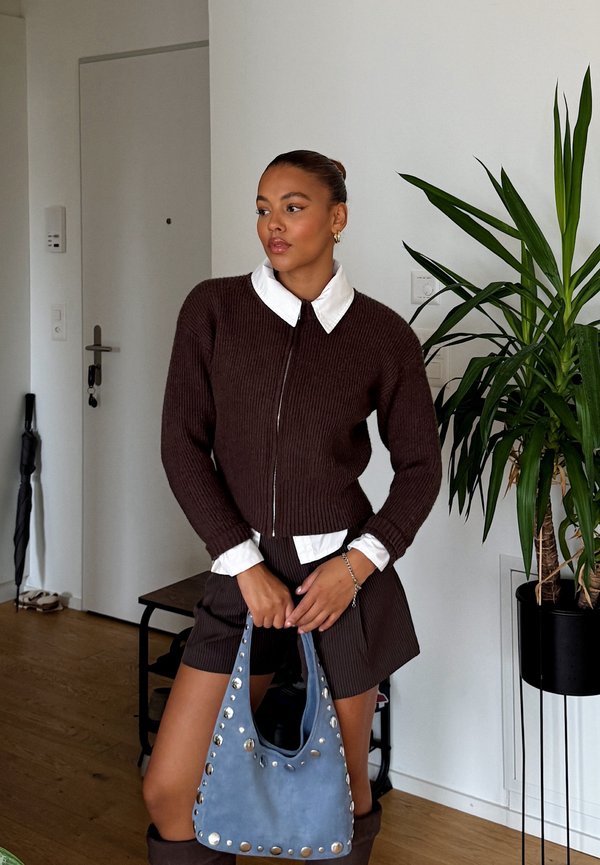 Ruskea ribattu villapaita, jossa on valkoinen kaulus, yhdistetty ruskeisiin shortsit. Kädessä vaaleansinisestä mokasta valmistettu laukku, jossa on niittikoristeita. Sisätila, jossa on viherkasveja.