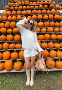 Žena u bijelom puloveru i čizmama drži bež torbu, stojeći ispred velikog drvenog izloga ispunjenog narančastim bundevama.