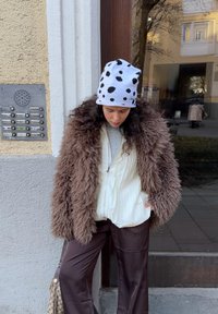 Manteau en fourrure brun duveteux sur une chemise blanche à fines rayures, assorti à un pantalon marron et un bonnet blanc à taches noires. Sac suspendu sur le côté.