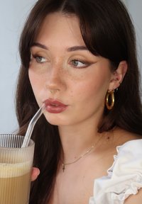 Transparent sugerør i et klart, ribbet glass fylt med en lys brun drikke. Personen har langt brunt h�år, fregner og bruker gullsmykker.