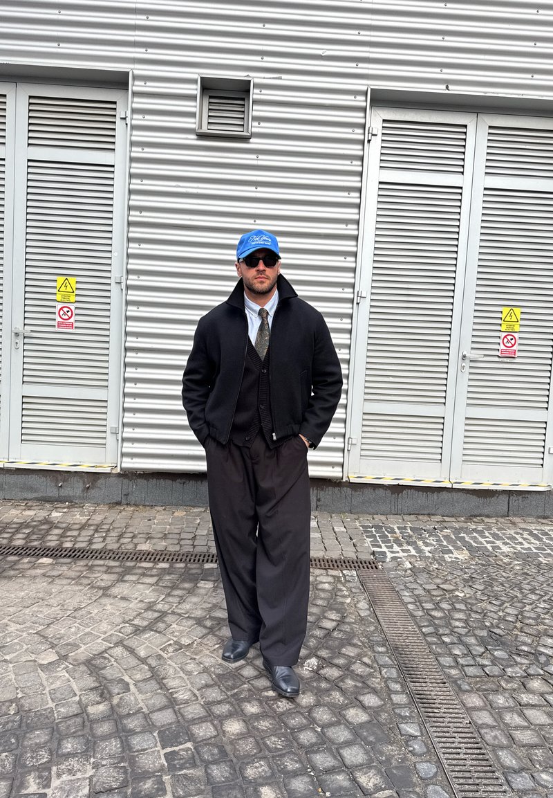 Homme portant un manteau noir, une cravate, un pantalon large noir, une casquette bleue et des lunettes de soleil, debout sur une rue pavée devant des portes métalliques grises.