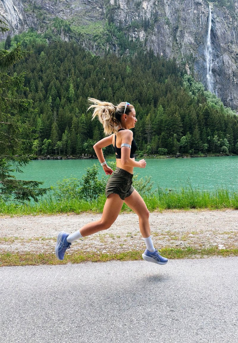Young woman in athletic wear running on a road by a lake, with forested mountains and a waterfall in the background.