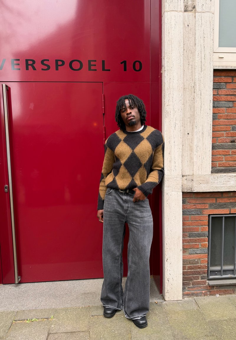 Homme avec dreadlocks portant un pull à motif losanges marron et noir et un jean large gris, debout devant une porte rouge numérotée 10.