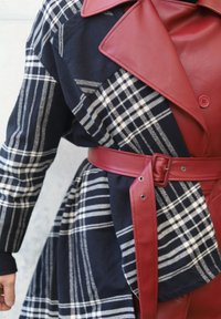 Crvena kaput ukrašen kožom s uzorkom mornarsko plave i bijele kariranosti, dizajniran s pojasom u struku i širokim ovratnikom, prikazujući slojevite materijale.