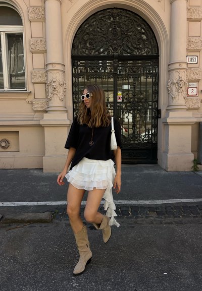 Mujer con blusa negra, falda blanca de varias capas, botas beige y gafas de sol blancas caminando frente a una elaborada reja negra de hierro forjado y un edificio beige.