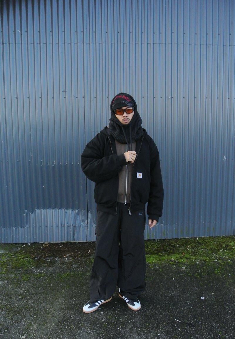 Μαύρο μπόμπερ μπουφάν πάνω από καφέ φούτερ, συνδυασμένο με φαρδιά μαύρα παντελόνια. Τα αξεσουάρ περιλαμβάνουν ένα μαύρο καπέλο και γυαλιά ηλίου με πορτοκαλί απόχρωση.