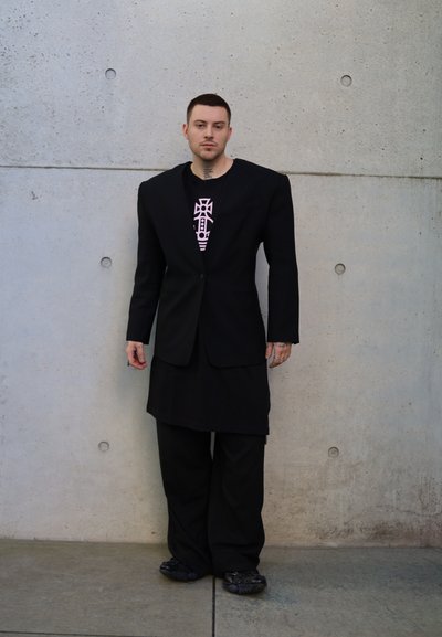 Blazer negro de gran tamaño sobre una camiseta negra con un gráfico en rosa, combinado con pantalones anchos negros y zapatillas negras. Fondo de concreto texturizado.