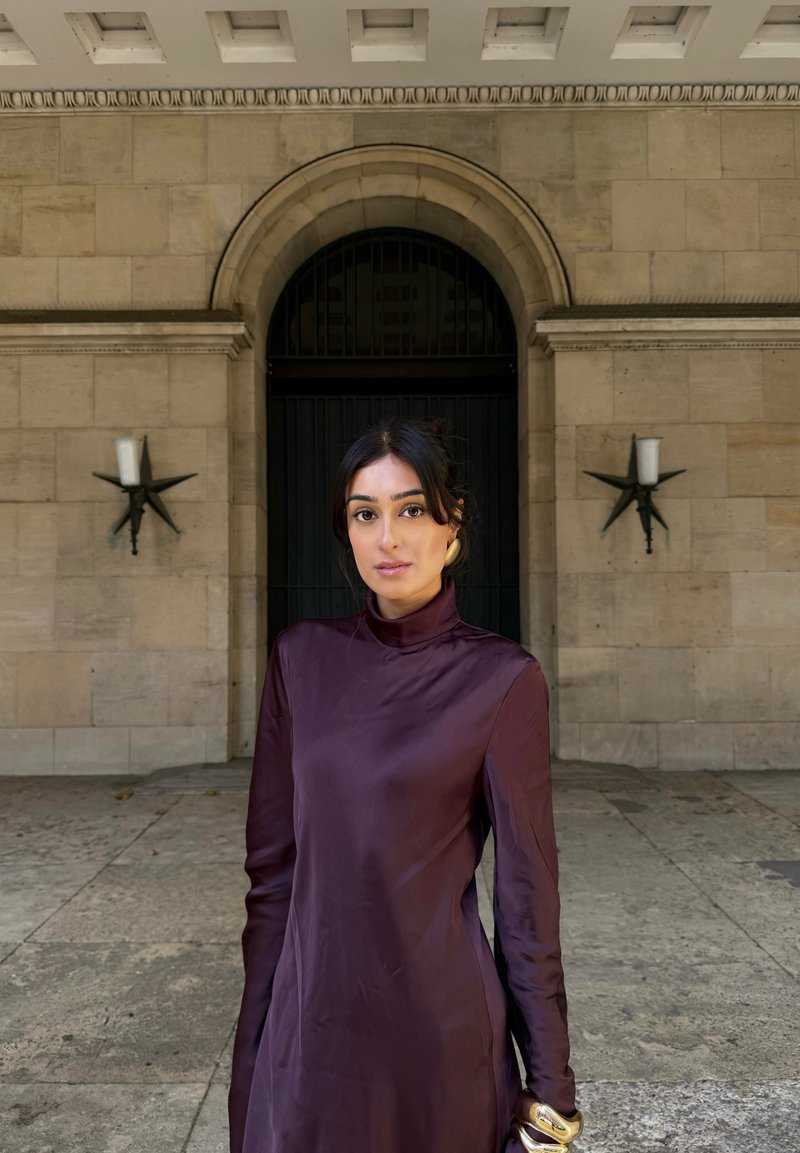 Abito a maniche lunghe con collo alto in raso viola scuro. Tessuto liscio, silhouette aderente. Bracciale d'oro al polso. Muro di pietra e appliques a forma di stelle sullo sfondo.