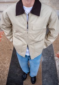 Veste en toile beige avec un col brun foncé, fermeture éclair et deux poches avant. Portée sur une chemise blanche avec un jean bleu clair et des chaussures noires.