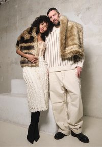 Duas modelos usam roupa exterior texturizada. Uma delas usa um xale em pelos sintéticos com riscas; a outra, um suéter oversized bege. A modelo feminina exibe uma saia creme com franjas.