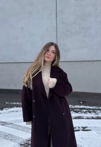 Νεαρή γυναίκα με μακριά μαλλιά, φορούσα σκούρο παλτό και μπεζ πουλόβερ, στέκεται σε χιονισμένο έδαφος μπροστά σε γκρι τσιμεντένιο τοίχο, κοιτώντας προς τα πλάγια.